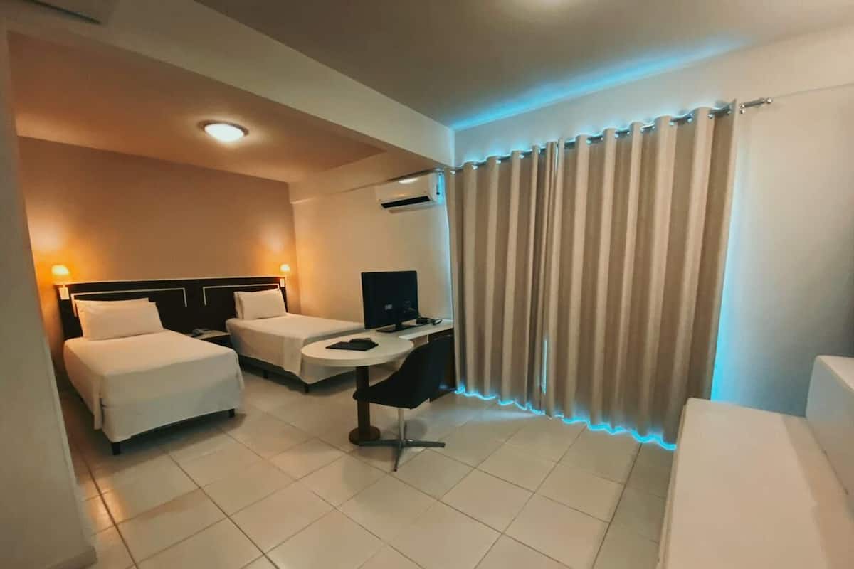 A hotel room with two beds, a desk, a chair, a TV, and a window with curtains.