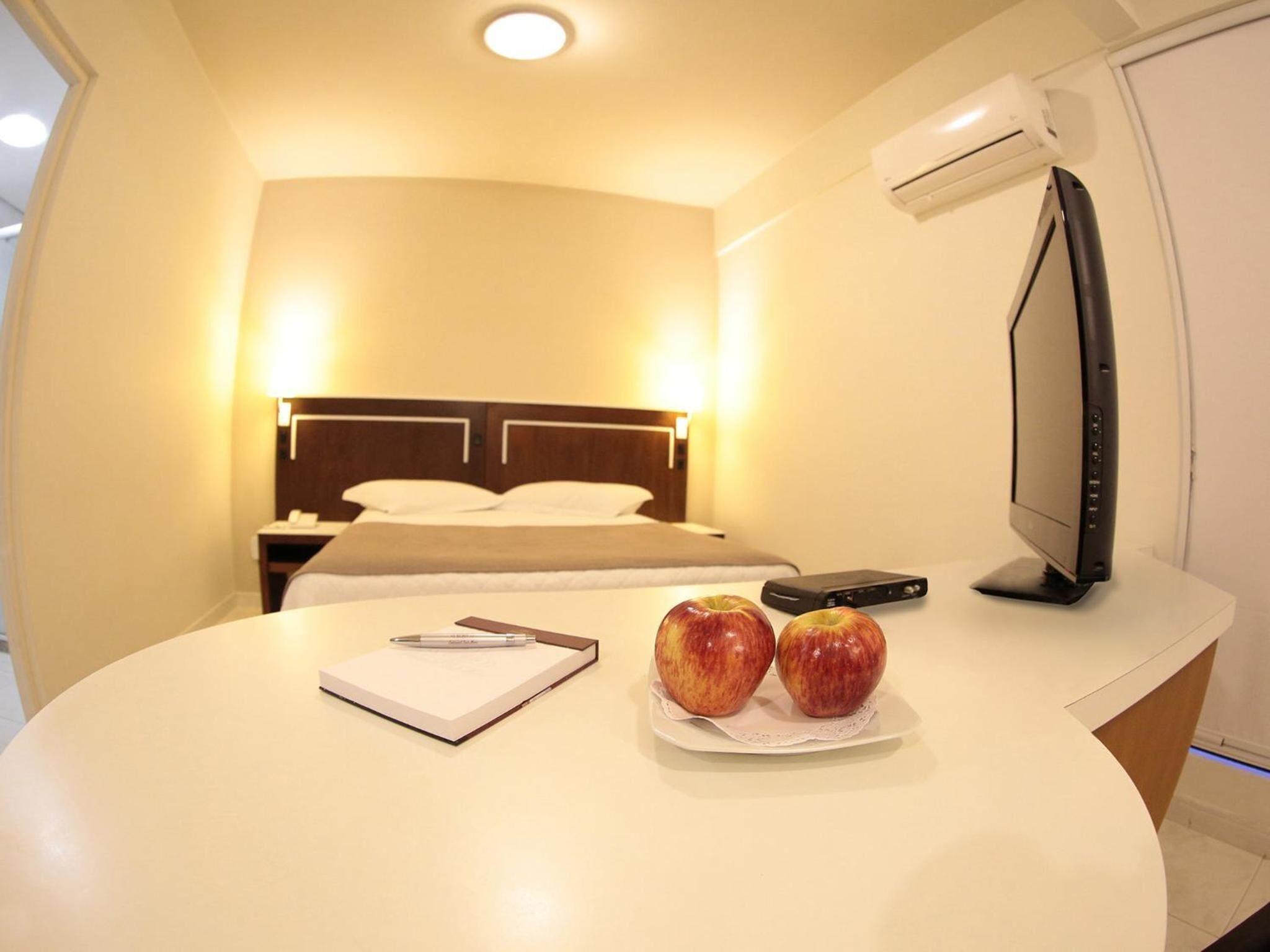 Ein Hotelzimmer mit Bett, Nachttisch und Schreibtisch, auf dem sich ein Computerbildschirm, ein Notizbuch und zwei Äpfel befinden.