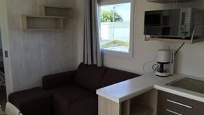 A compact living space with a sofa, a small kitchenette, and a window with curtains. - Camping Pen Guen (Saint-Cast-le-Guildo)