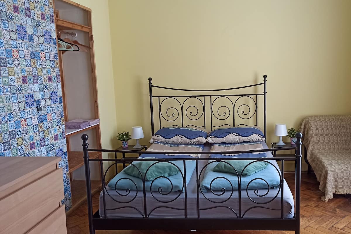 Ein Bett mit Metallgestell, zwei Nachttische mit Lampen, eine Wand mit Muster, eine Holzkommode und ein Sofa mit gemustertem Bezug.