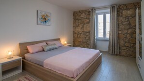 A bedroom with a wooden bed, a nightstand with a lamp, a stone wall, and a window with curtains. - Apartment for 4 person (Vrbnik)