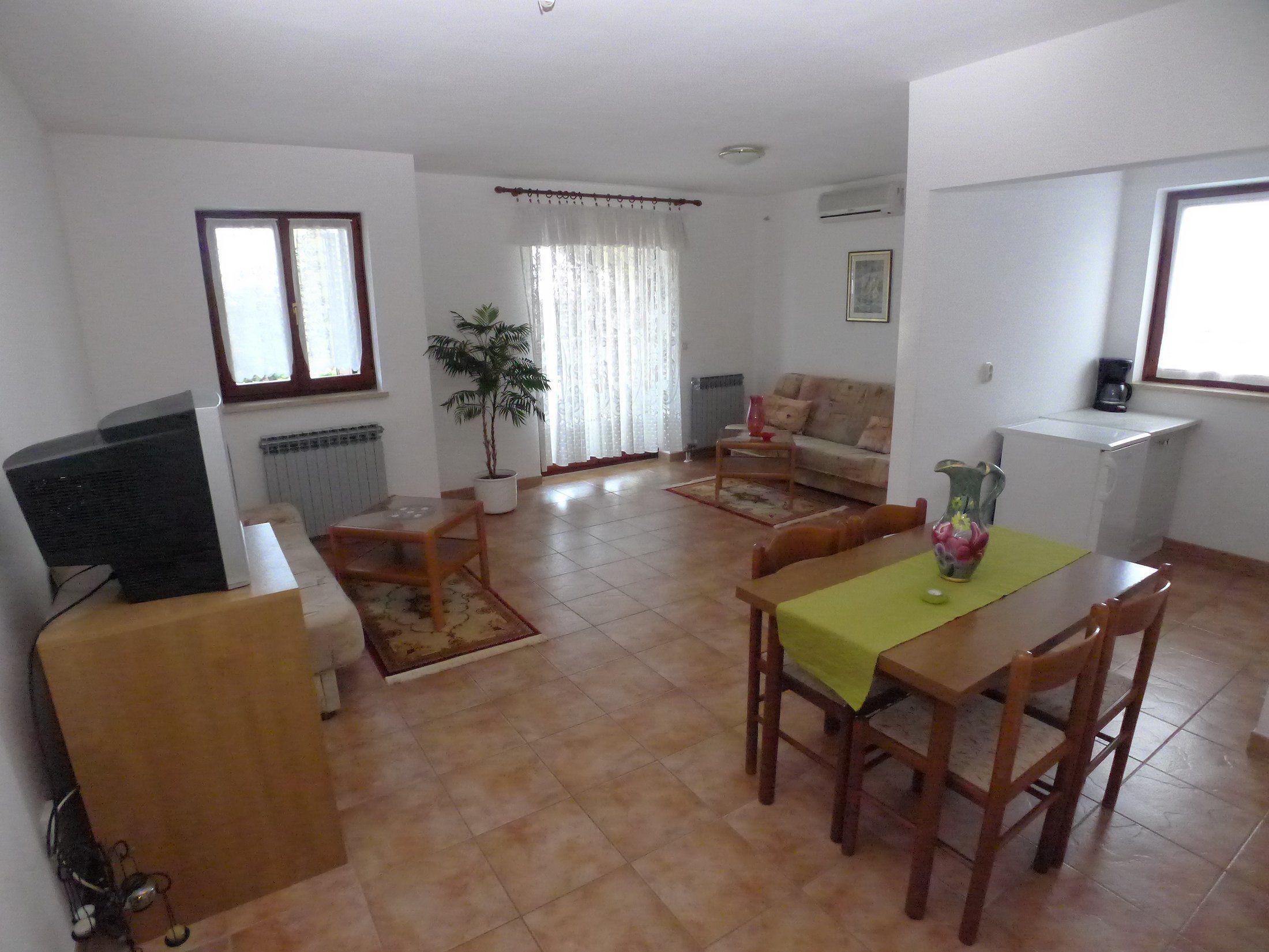 Uma sala de estar com sofá, mesa de jantar com cadeiras, televisão, radiador, planta em vaso e janela com cortinas.