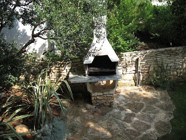 Enjoy al fresco dining in a serene garden setting with a stone-built barbecue grill and lush greenery.