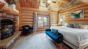 A log cabin bedroom with a fireplace, a bed, a blue sofa, and a ceiling fan. - Highlands Treehouse with Shuffleboard & Private Hot Tub! Golf Course & Mountain Views! (Breckenridge)