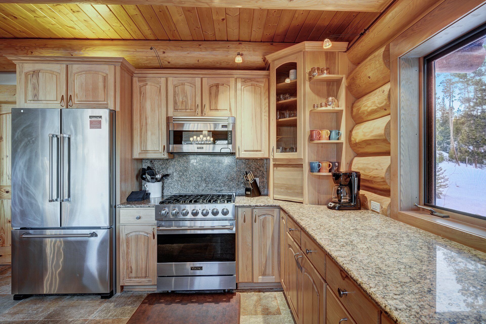 Cocina de cabaña rústica con electrodomésticos de acero inoxidable, armarios de madera y un ventanal con vistas al exterior.