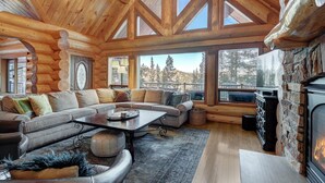 A log cabin interior with a large sectional sofa, a fireplace, and large windows offering a view of the surrounding forest. - Highlands Treehouse with Shuffleboard & Private Hot Tub! Golf Course & Mountain Views! (Breckenridge)