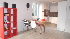 Eine moderne Küche mit weißen Schränken, ein Essbereich mit Holztisch und -stühlen sowie ein rotes Bücherregal.