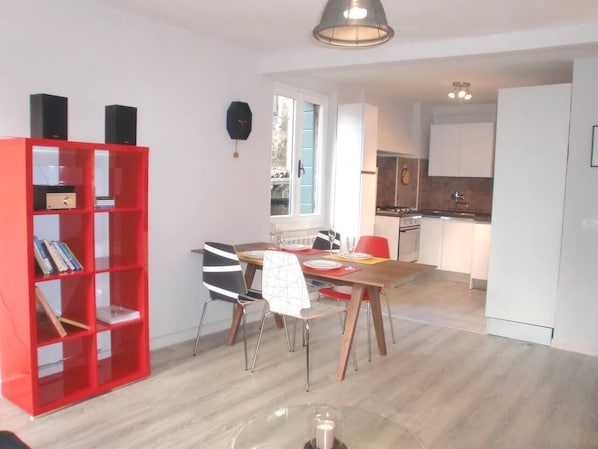A modern kitchen with white cabinets, a dining area with a wooden table and chairs, and a red bookshelf.