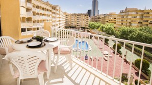 Un balcon avec des chaises blanches et une table, donnant sur une piscine et une cour intérieure agrémentée de verdure, avec des bâtiments en arrière-plan.
