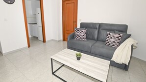 Un salon avec un canapé gris, une table basse et une petite plante. On y trouve une porte en bois et un mur blanc orné d’une horloge.
