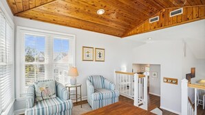 Une pièce avec un plafond en bois, une table à manger, deux fauteuils à rayures, une lampe et une vue sur une maison par la fenêtre.