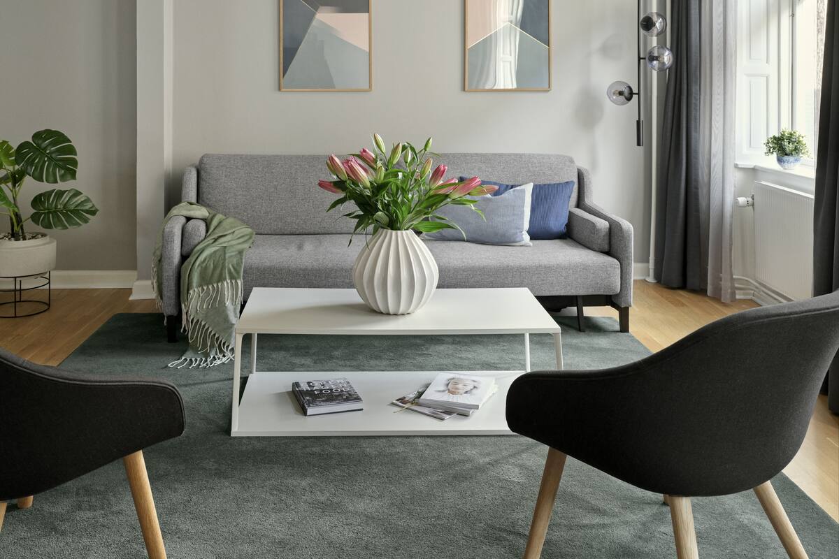 Un salon moderne avec un canapé gris, une table basse blanche et deux chaises noires. Deux tableaux abstraits ornent le mur, ainsi qu’une plante en pot et un vase rempli de fleurs sur la table basse.