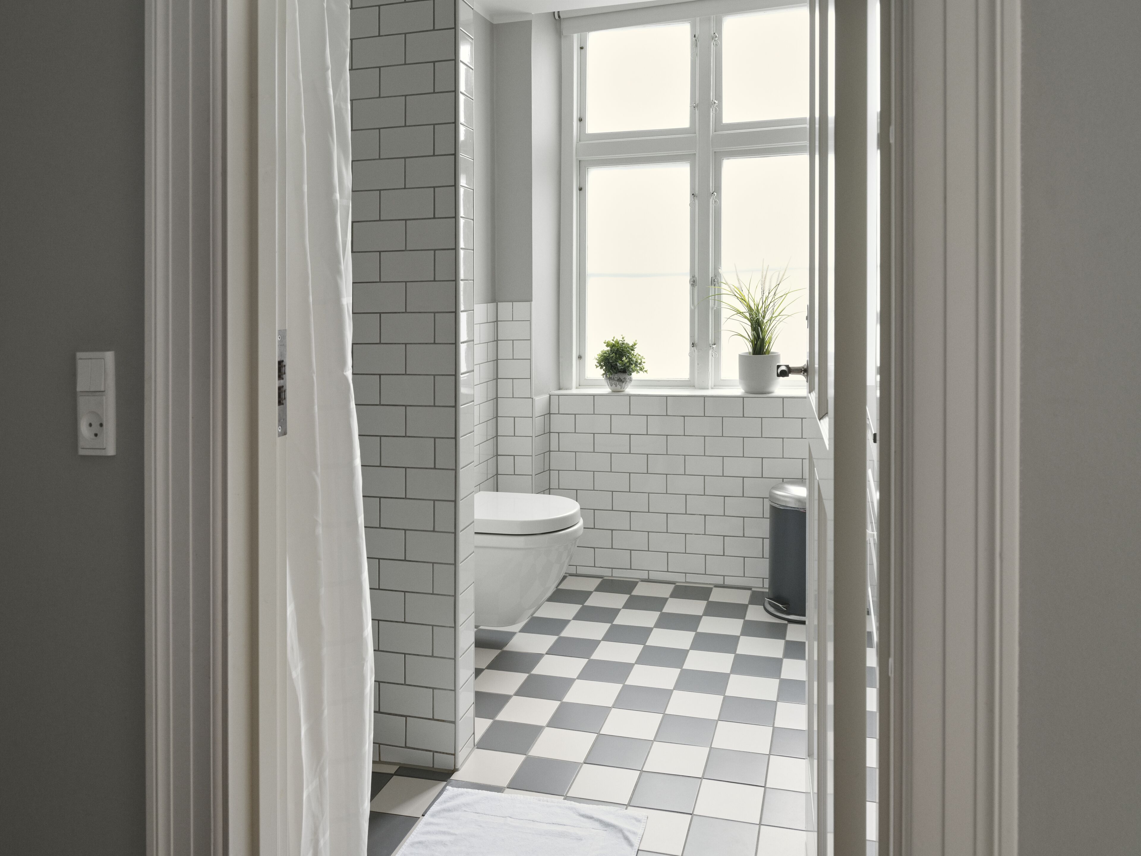 A bathroom with white tiled walls, a checkered floor, a toilet, a window with plants, and a visible electrical outlet.
