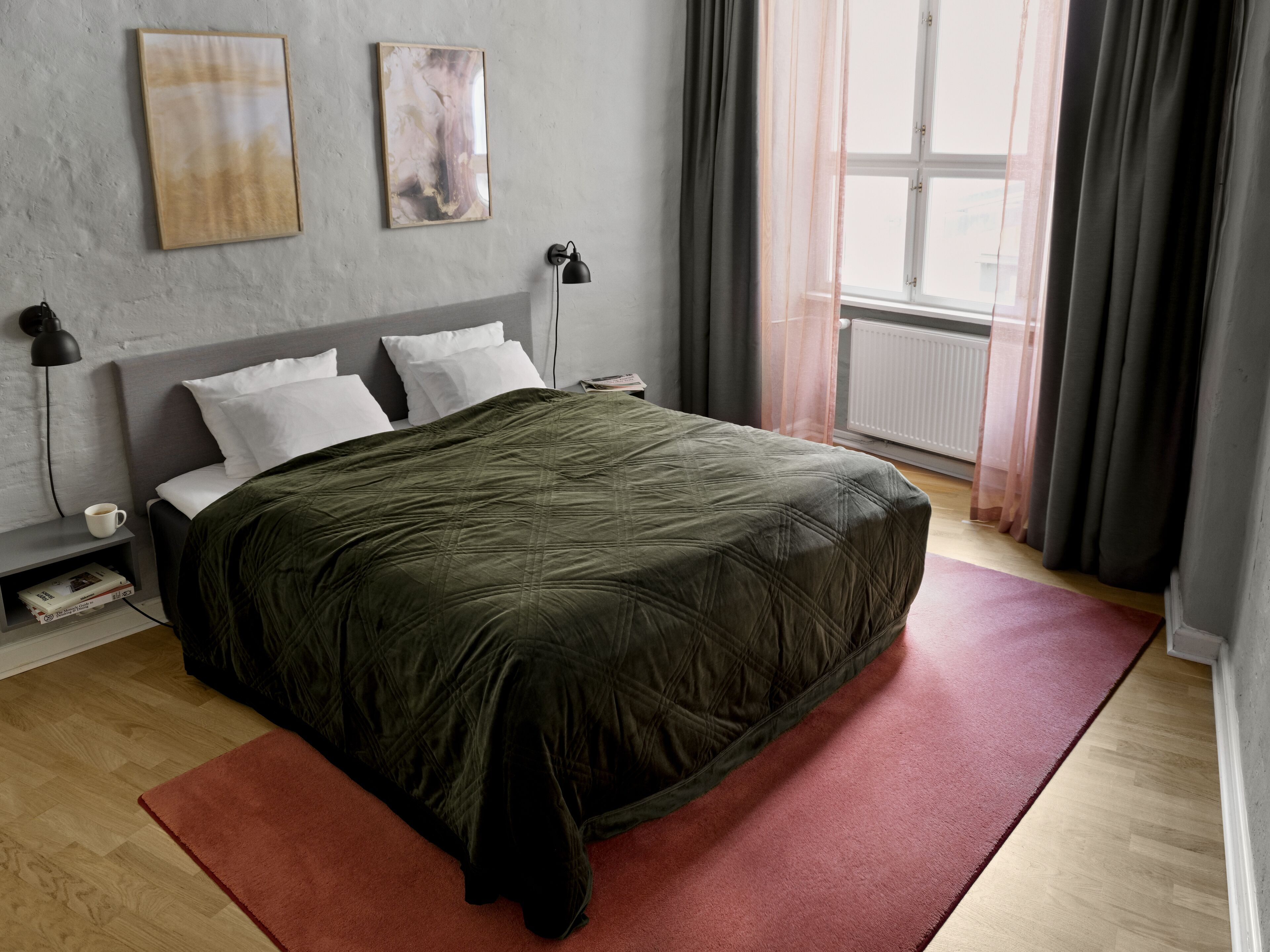 A neatly made bed with a dark green quilt, a red rug, and a grey headboard. There are two wall-mounted lamps and a framed abstract painting above the bed.