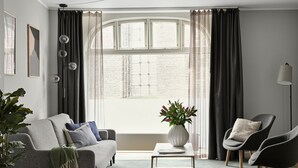 Una sala de estar moderna con un sofá gris, una mesa de centro y una silla. Hay una ventana con cortinas, una planta y un florero con flores.