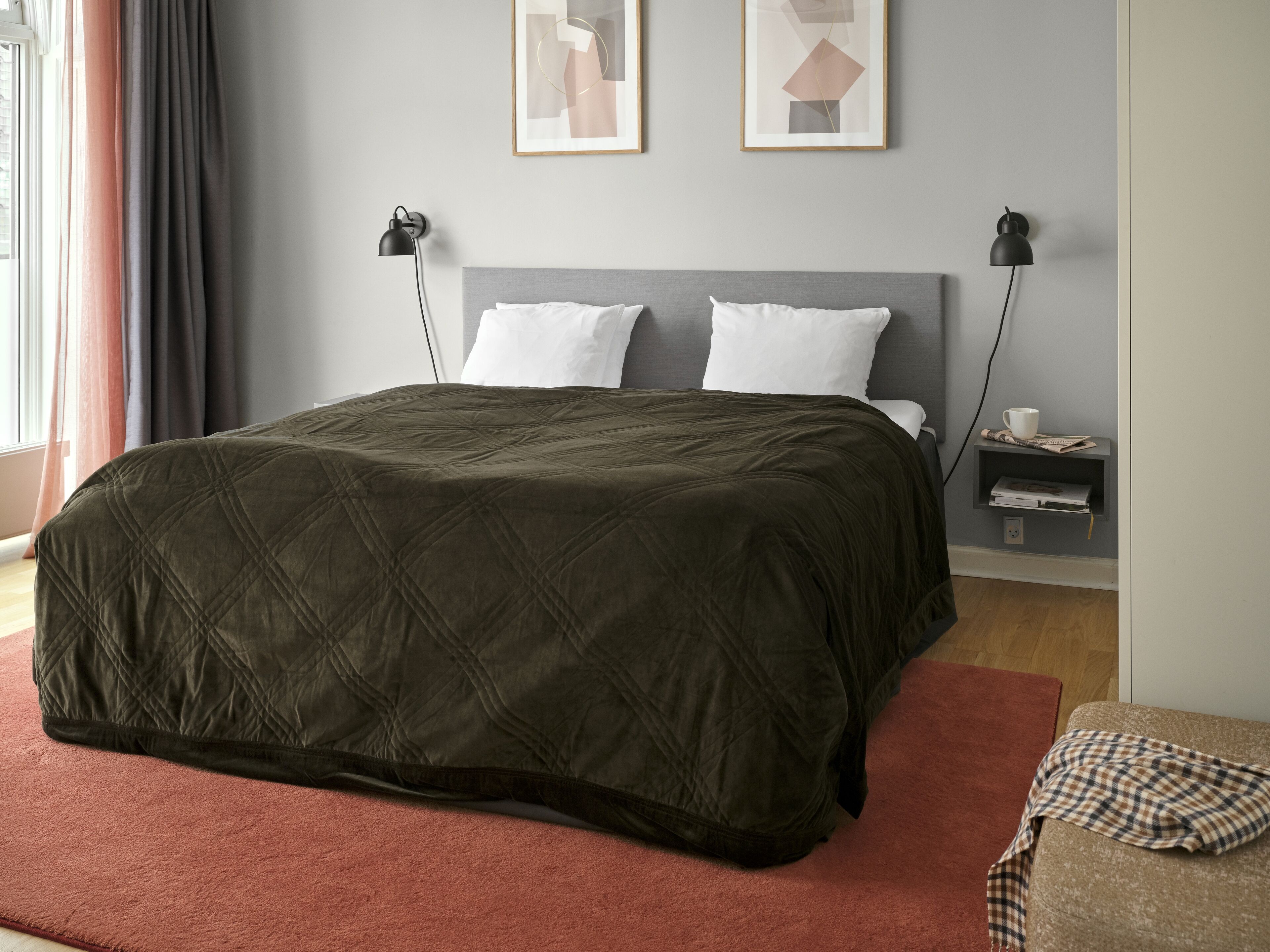 Una cama bien hecha con un edredón oscuro, dos lámparas de noche, una mesita auxiliar con una taza y un libro, y una obra de arte abstracto enmarcada en la pared.