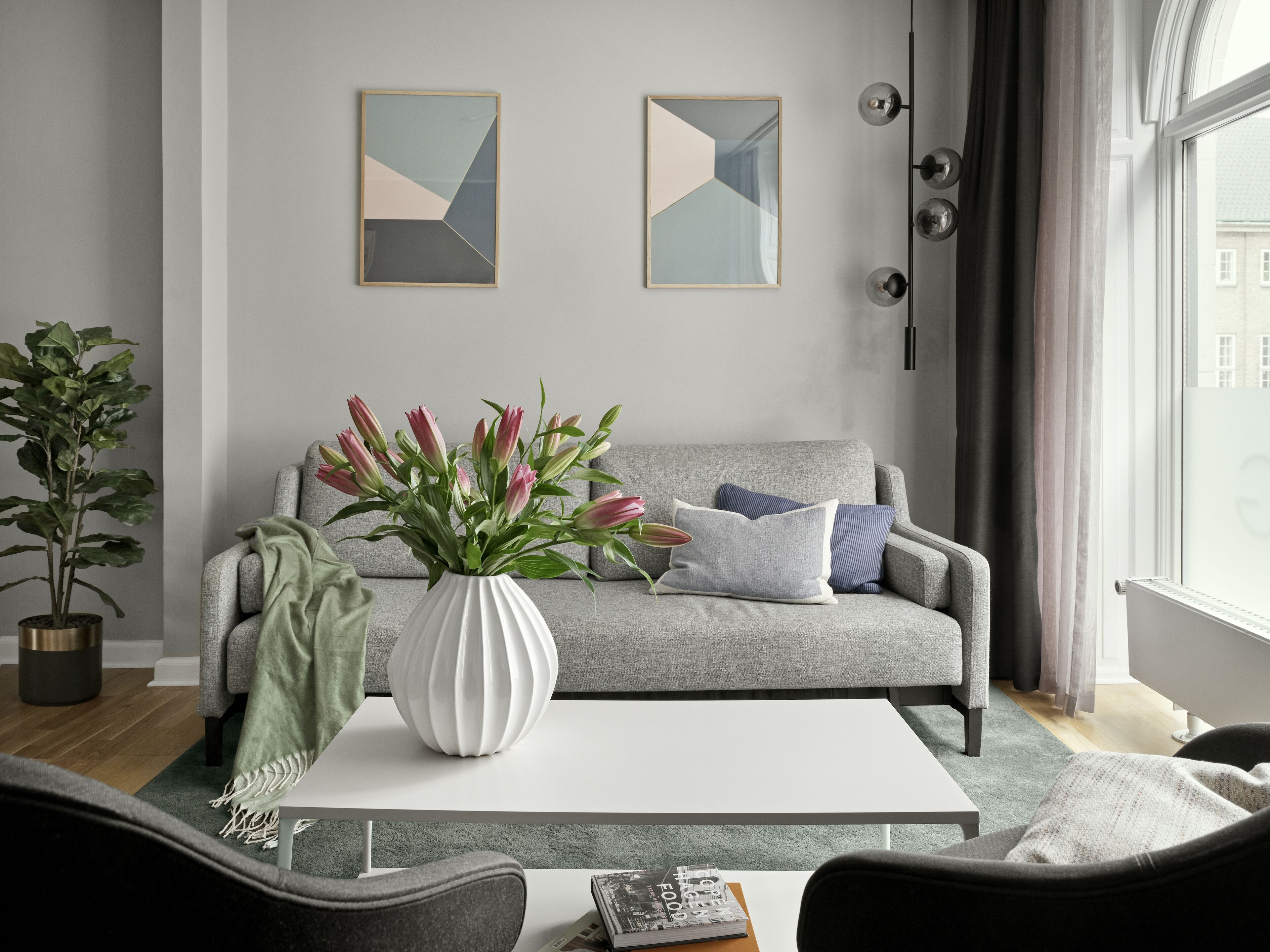 A modern living room with a grey sofa, a white coffee table, and a vase with flowers. There are two abstract paintings on the wall and a potted plant on the left.