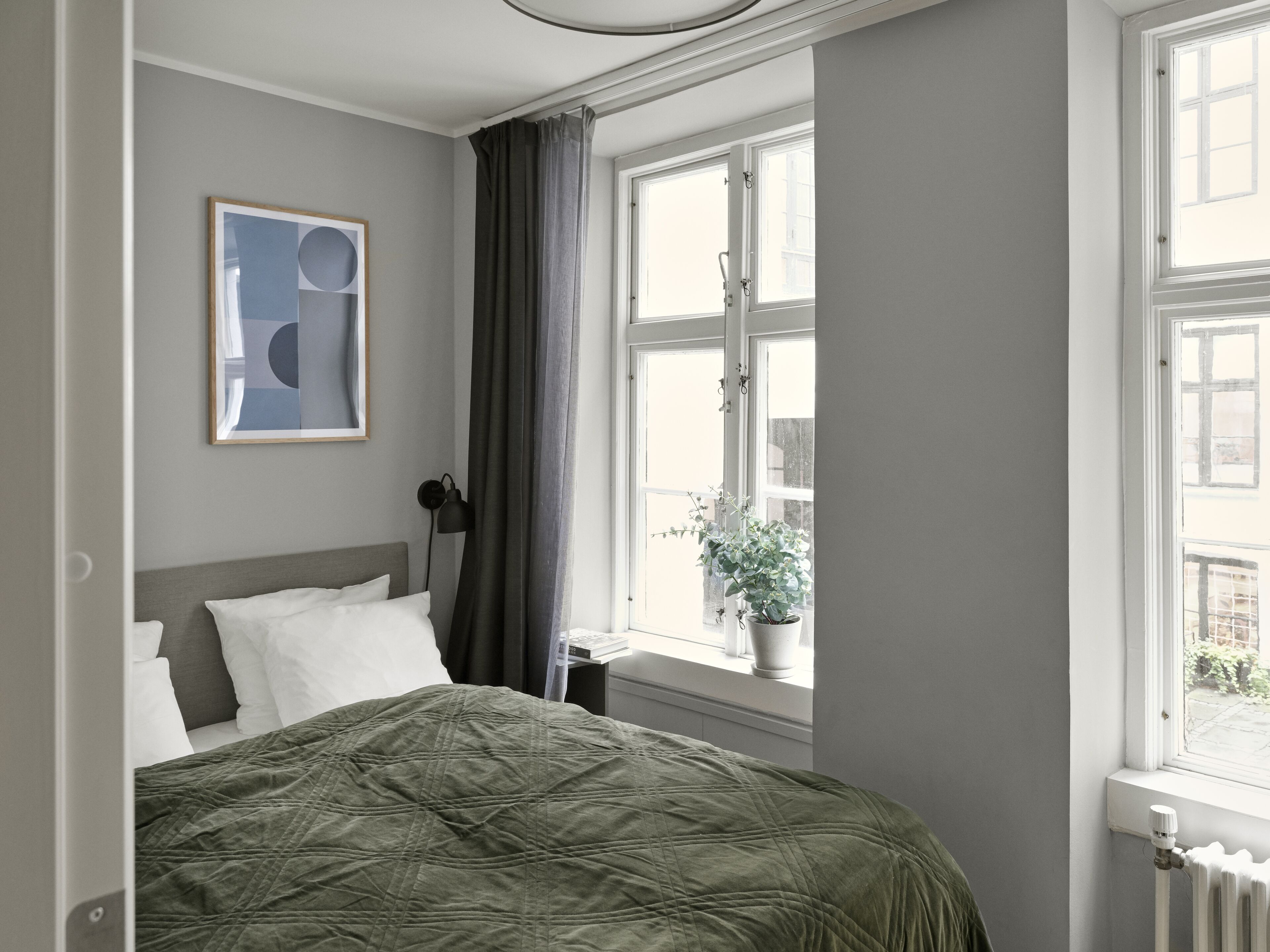 Ein Schlafzimmer mit Bett, einem Fenster mit Vorhängen, einem Heizkörper und einem gerahmten Bild an der Wand.