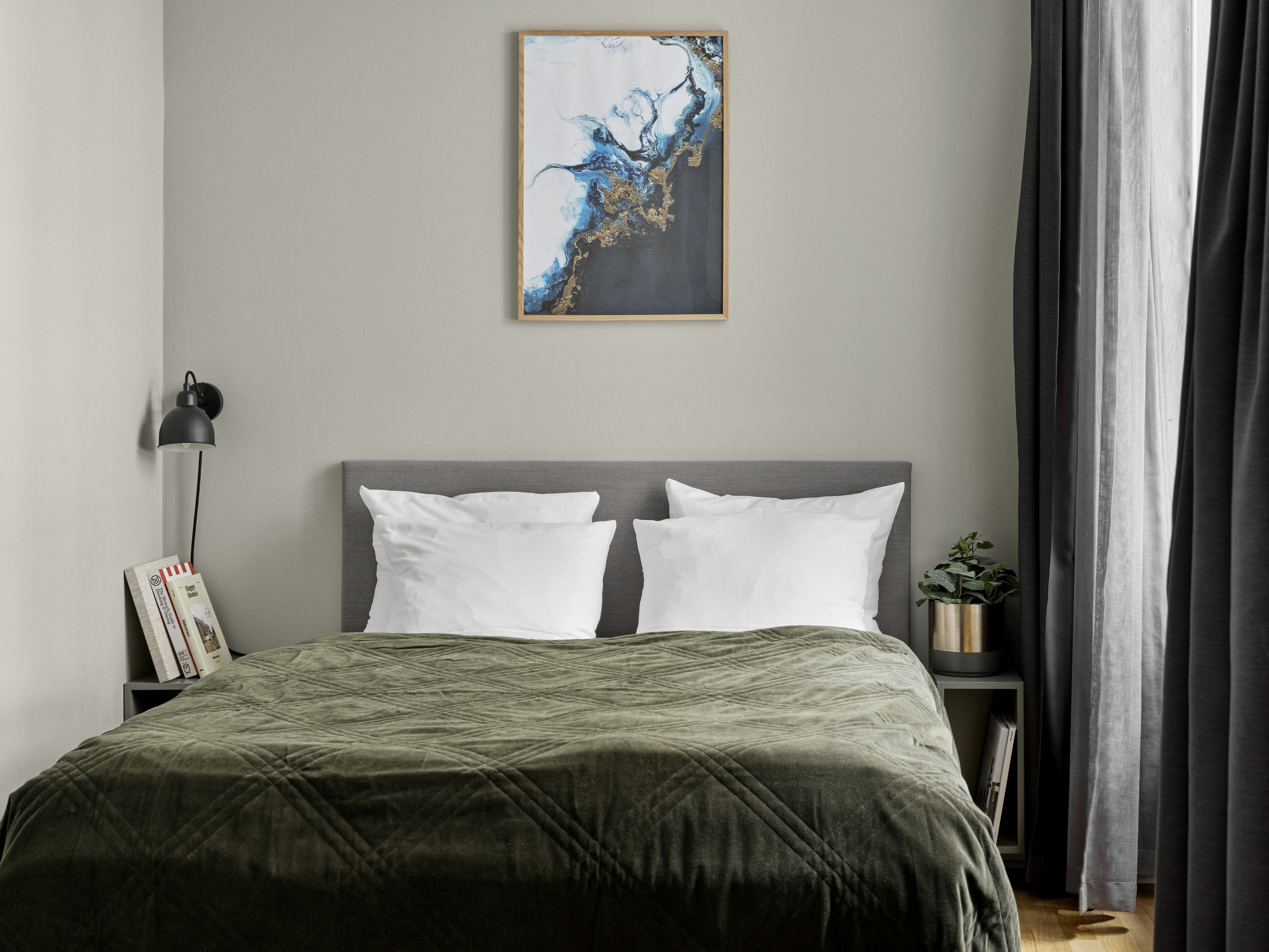 Ein modernes Schlafzimmer mit grauem Kopfteil, einer gesteppten grünen Bettdecke, zwei weißen Kissen, einem Nachttisch mit Lampe, einer kleinen Topfpflanze und einem gerahmten abstrakten Gemälde an der Wand.