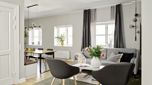 Una sala de estar moderna con un sofá gris, dos sillas negras, una mesa de comedor con sillas, un jarrón blanco con flores y una estantería.