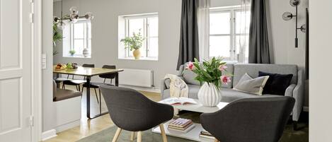 Een moderne woonkamer met een grijze bank, twee zwarte stoelen, een eettafel met stoelen, een witte vaas met bloemen en een boekenkast.