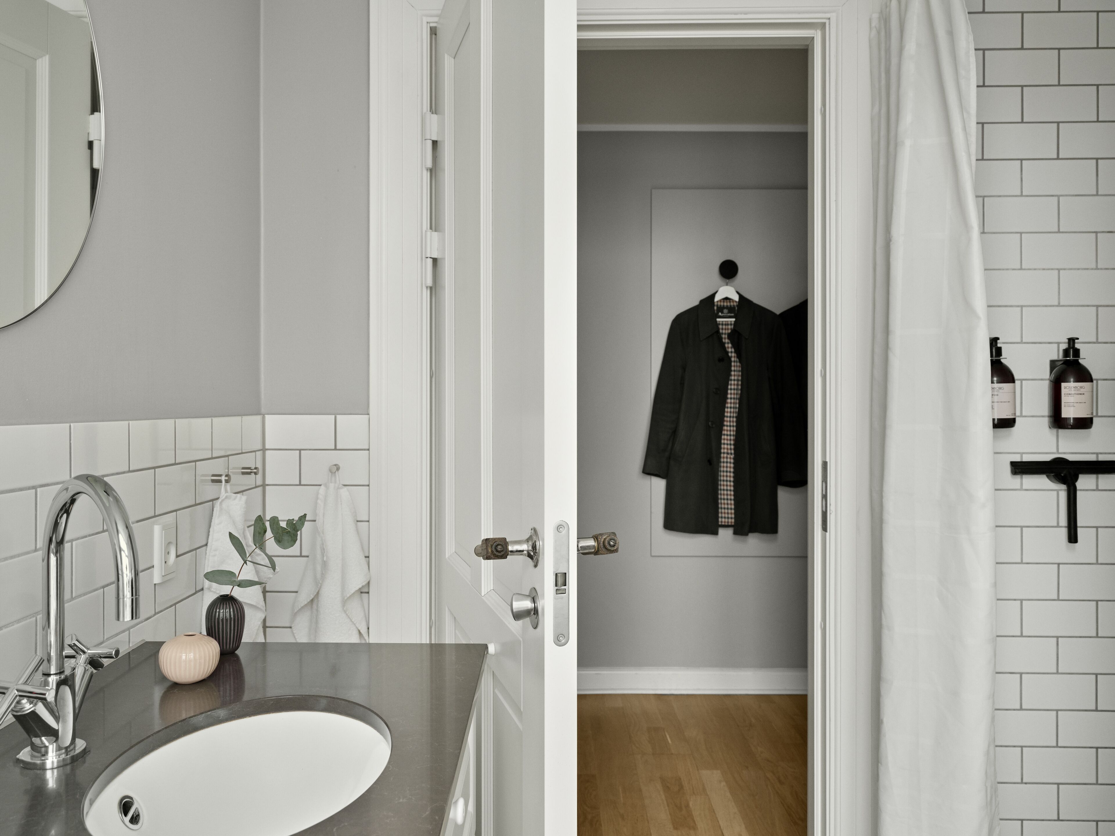 Un baño con lavamanos, espejo y barra para toallas. Hay una puerta que conduce a una habitación con perchero y artículos de aseo.