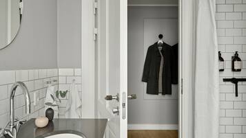 Un baño con lavamanos, espejo y barra para toallas. Hay una puerta que conduce a una habitación con perchero y artículos de aseo.