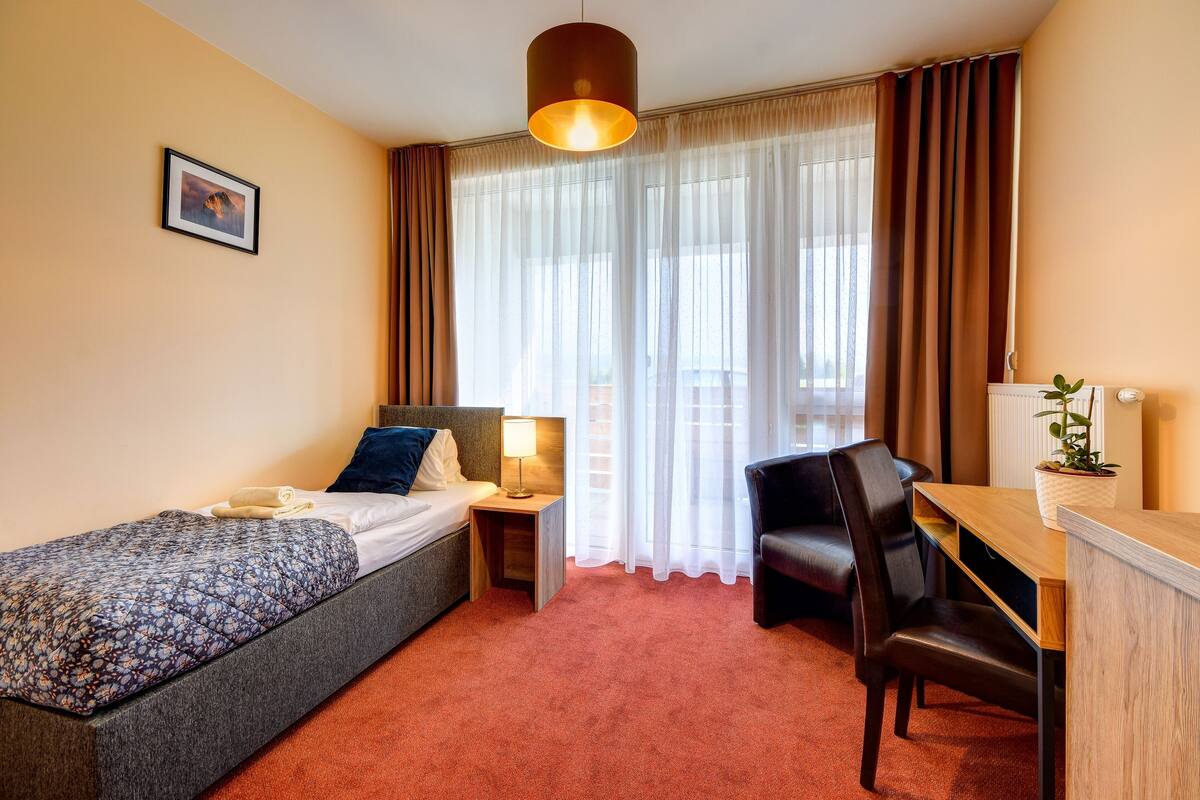 A hotel room with a single bed, a desk, a chair, a lamp, and a potted plant.