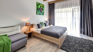 A modern bedroom with a wooden bed frame, a bedside table with a lamp, a green throw on the sofa, and a large window with curtains. - Penzión Čerešnový Sad (Mengusovce)