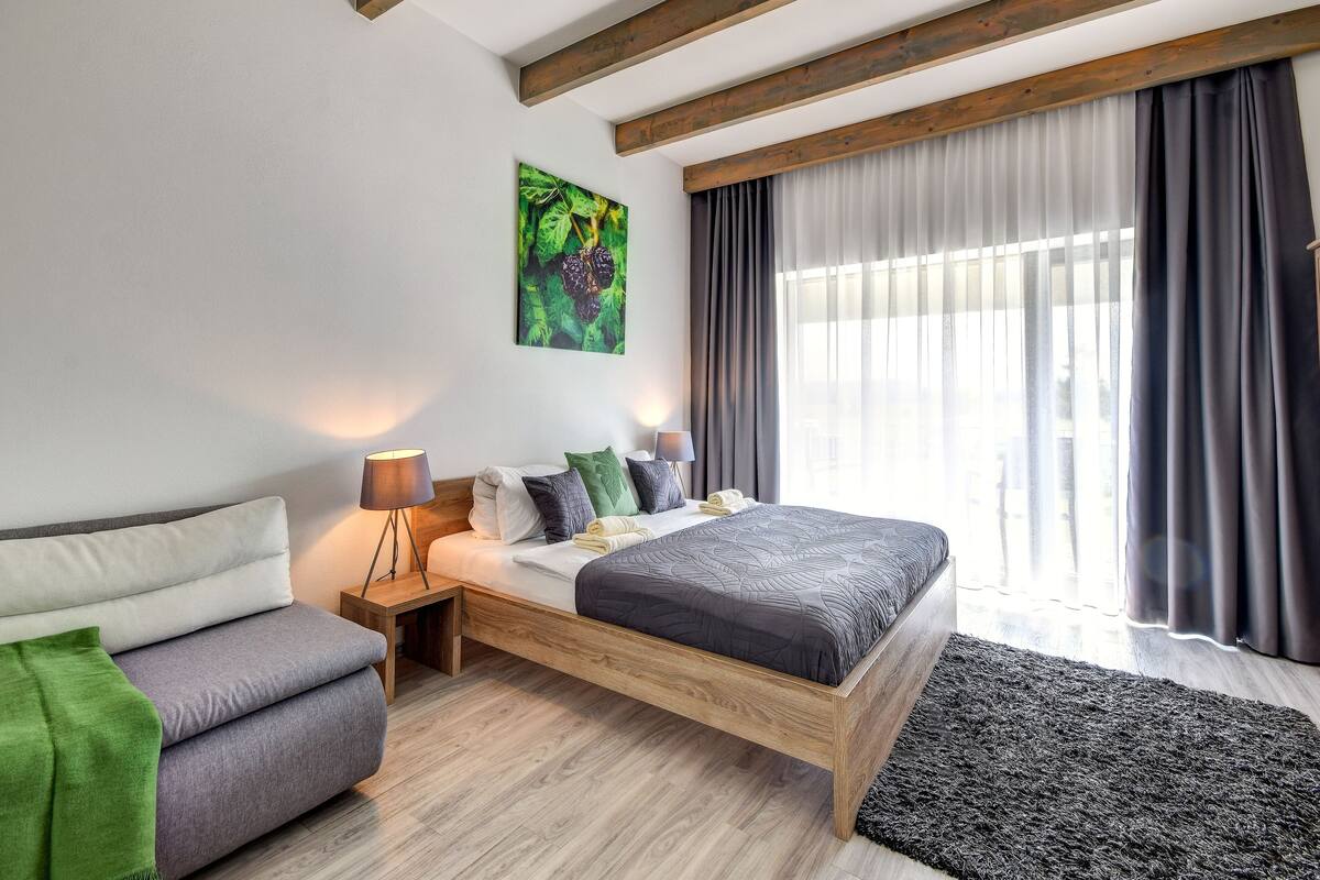 A modern bedroom with a wooden bed frame, a bedside table with a lamp, a green throw on the sofa, and a large window with curtains.