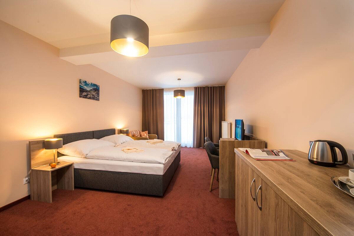 A hotel room with a bed, bedside table, lamp, desk, chair, and a view of the outdoors.