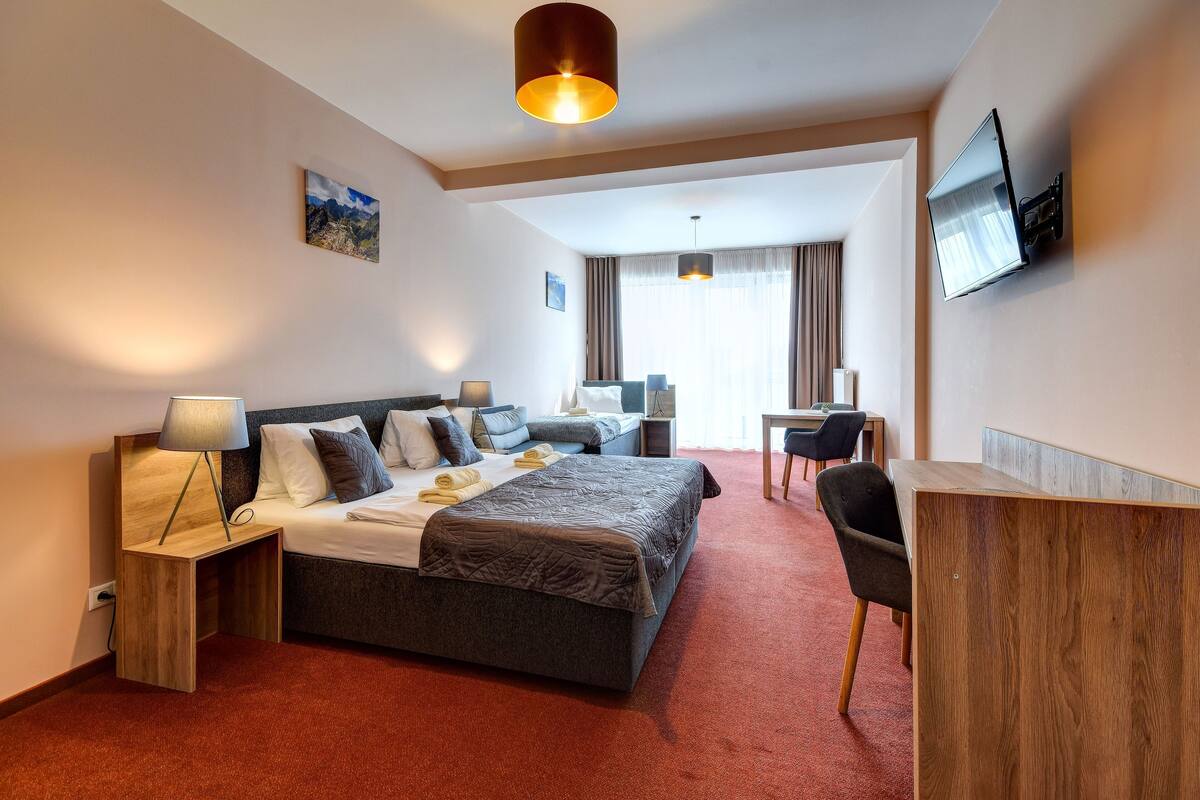 A hotel room with a large bed, bedside tables, a desk, a chair, a TV, and a window with curtains.