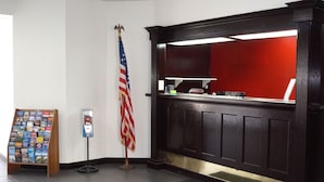 A reception area with a wooden desk, a red backdrop, an American flag, and a travel brochure stand. - Super 7 Inn (Midland)