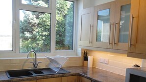 A kitchen with wooden cabinets, a washing machine, a microwave, and a window with a view of trees. - Filton House (Bristol)