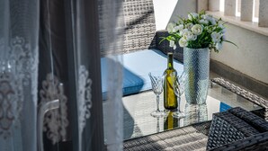 Een balkon met rieten meubels, een glazen tafel, een vaas met witte bloemen en een fles wijn.