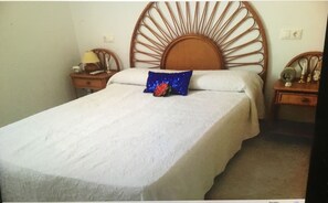 Una cama con cabecera de madera, una colcha blanca y una almohada decorativa azul. Hay dos mesitas de noche con lámparas y un reloj.