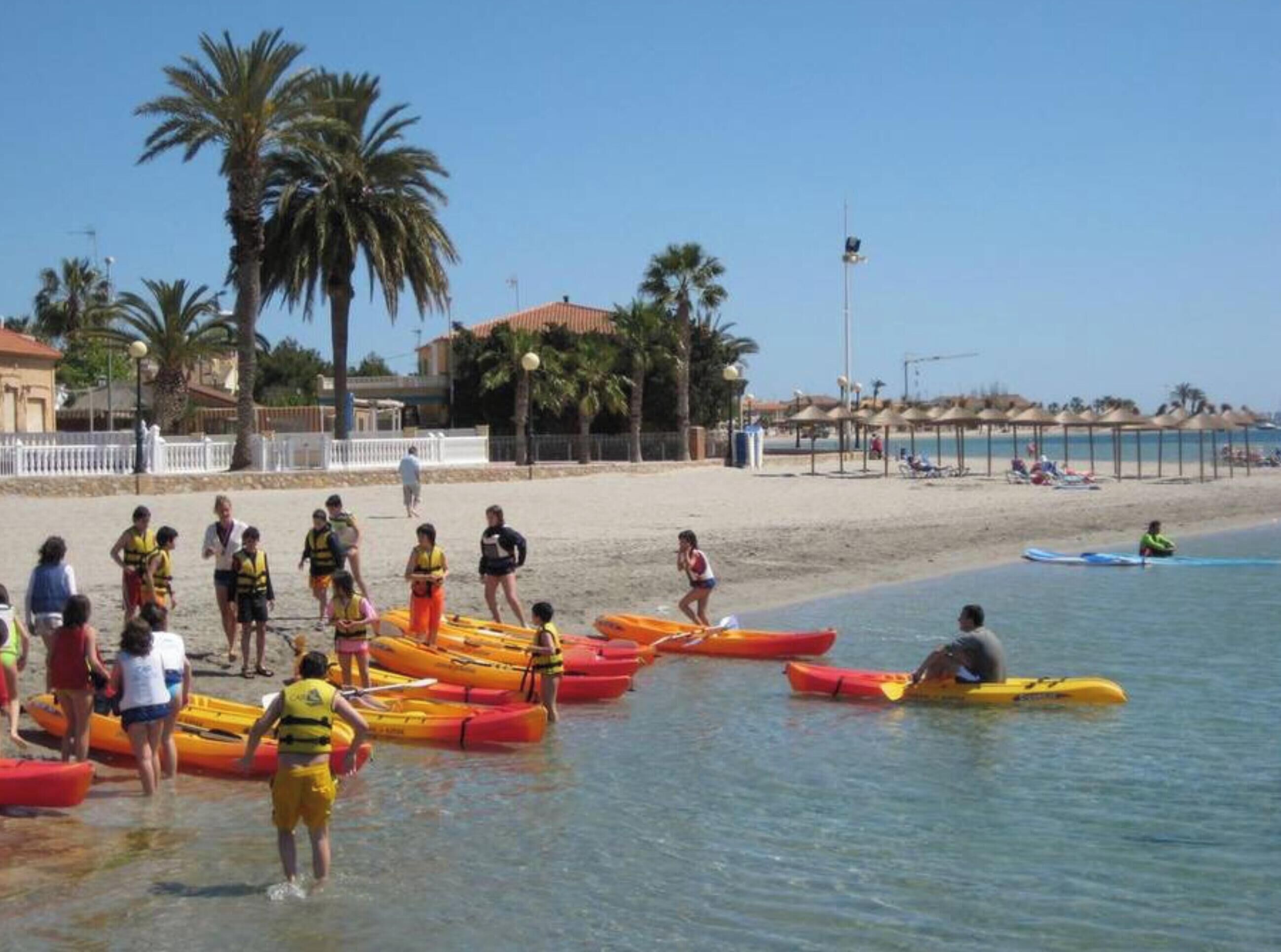 A beach with people in kayaks and life jackets, palm trees, and beach umbrellas.