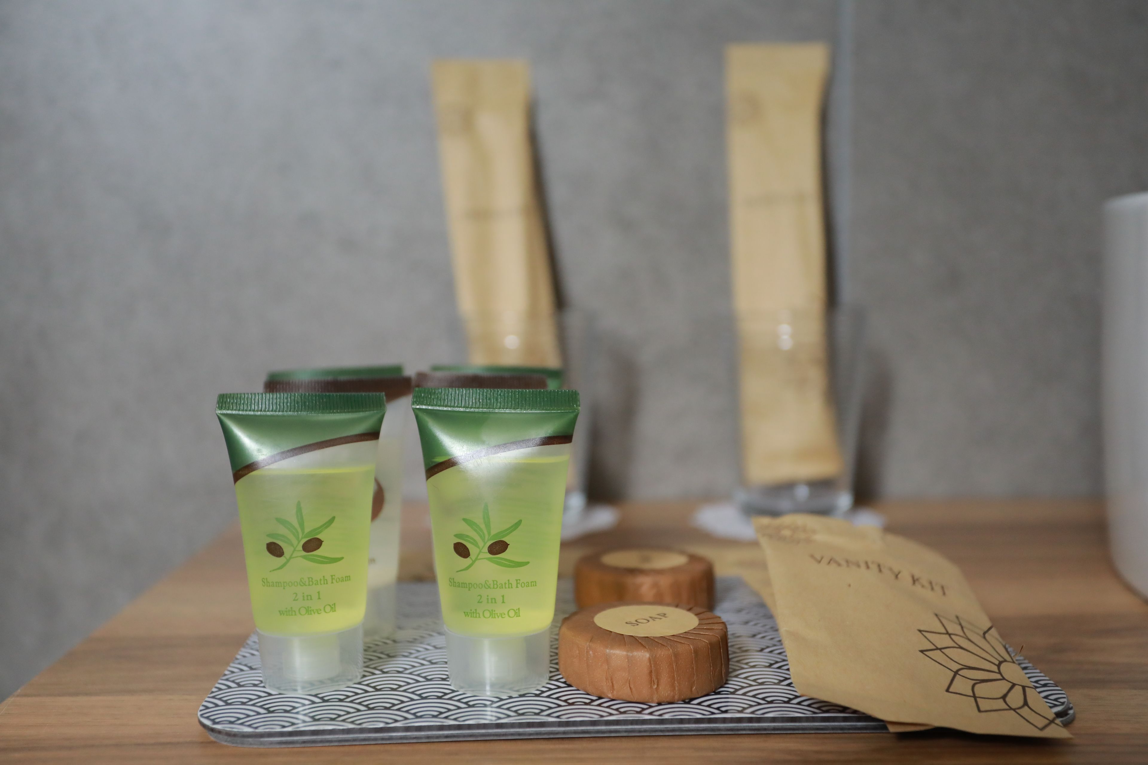 Two tubes of shampoo and bath foam with olive oil, a wooden soap bar, and a vanity kit bag on a patterned tray.