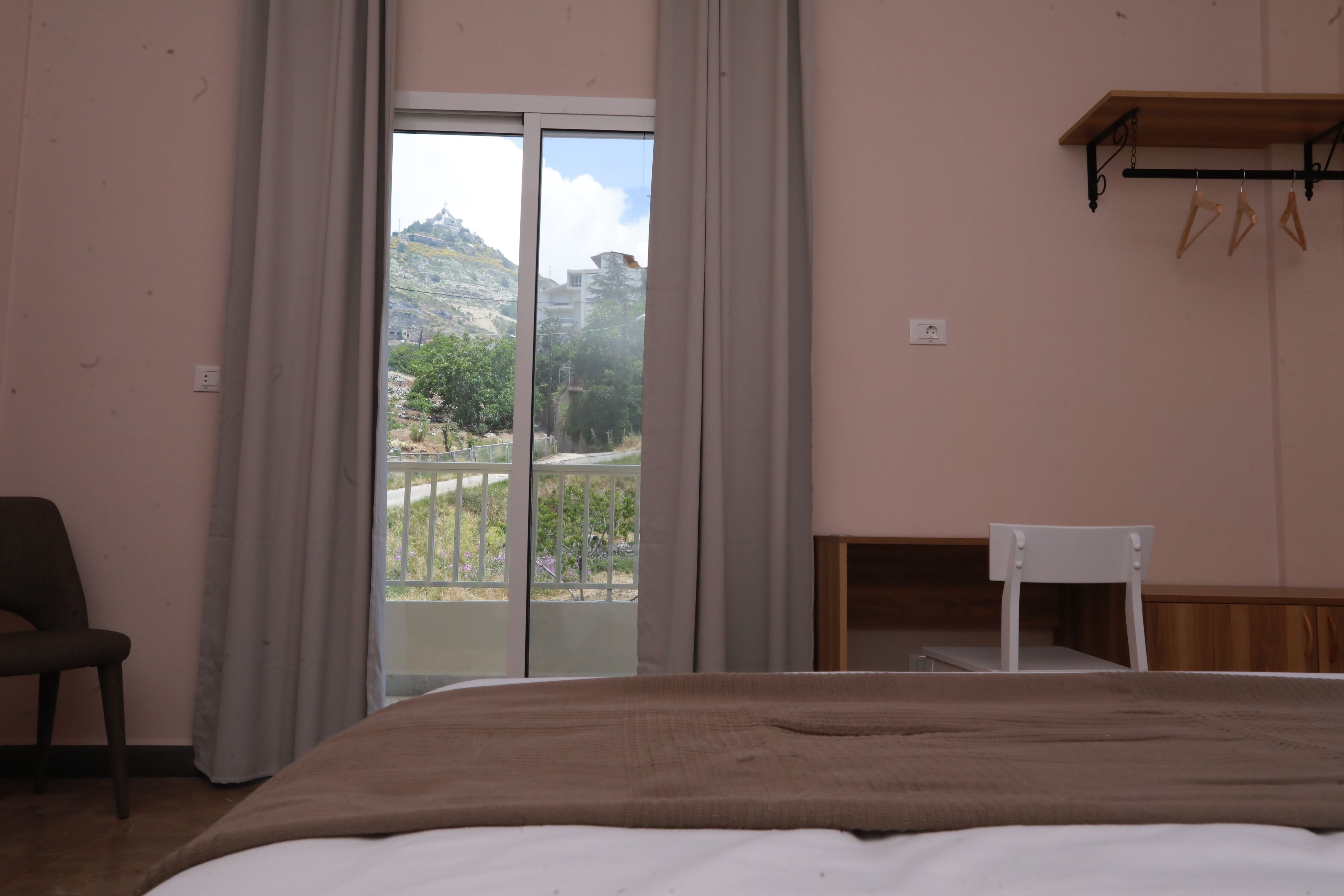 A bedroom with a bed, a chair, a wooden shelf, and a view of a balcony with a mountain in the background.