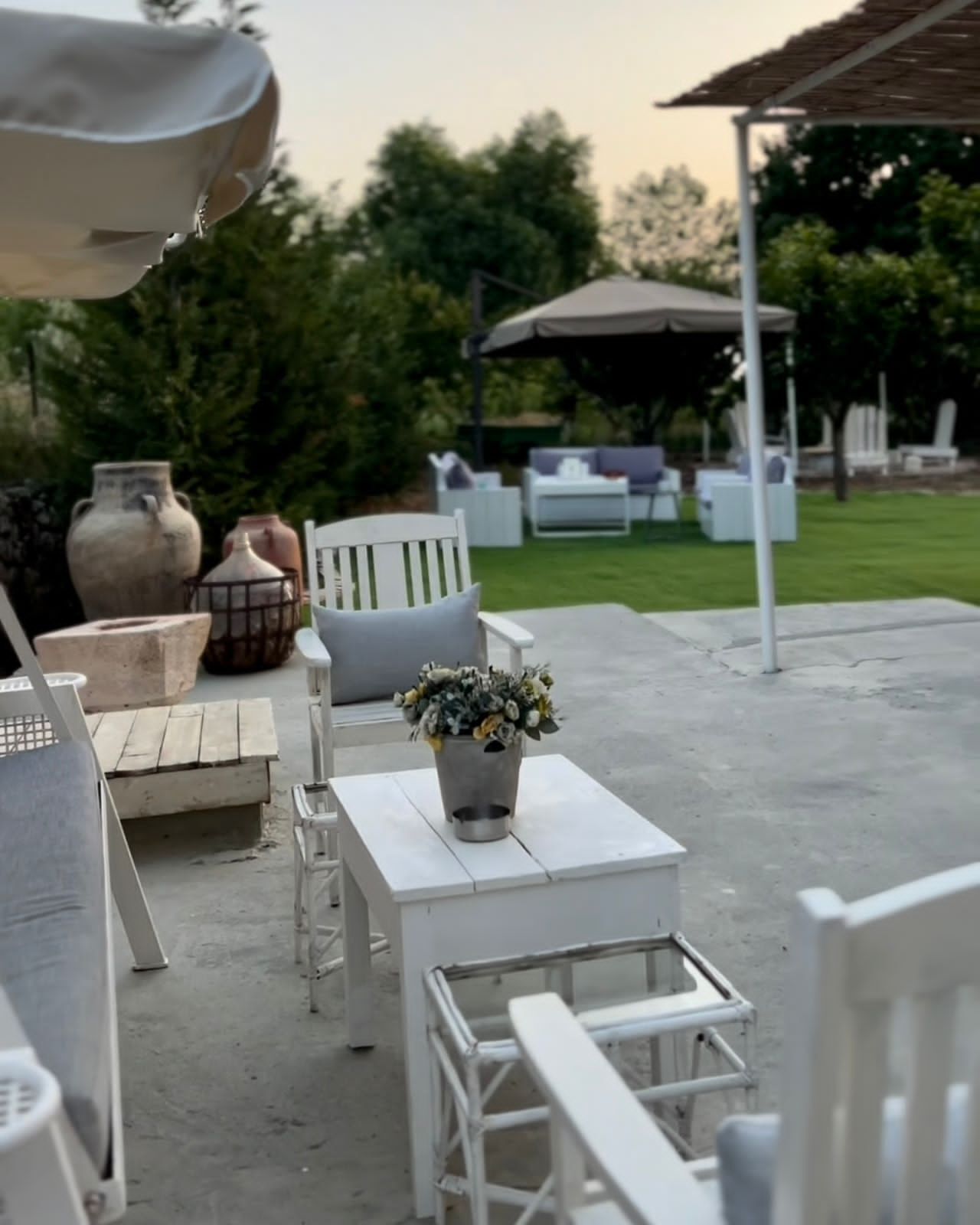 An outdoor seating area with white furniture, a metal table with a flower arrangement, and a pergola in the background.