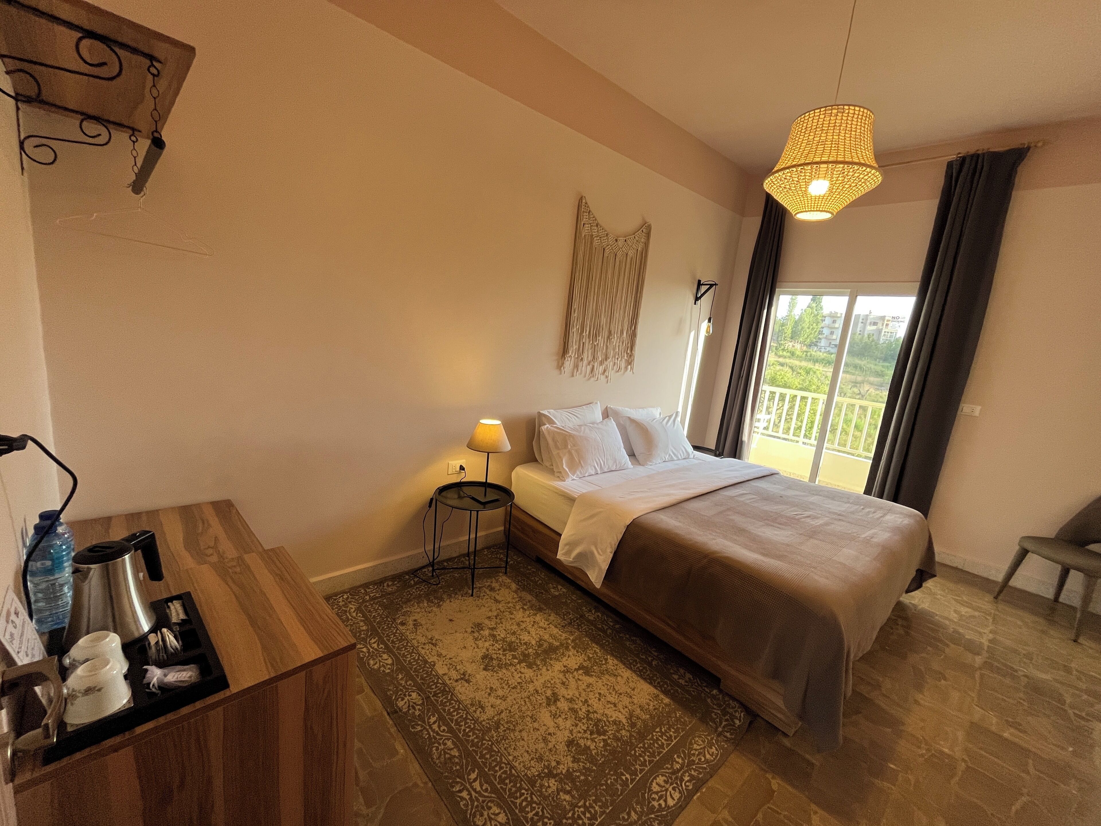 A hotel room with a bed, bedside table, lamp, and a view of the outdoors.