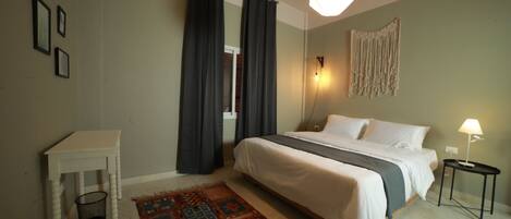 A bedroom with a bed, a bedside table, a small table, a floor lamp, a wall-mounted light, and a patterned rug.