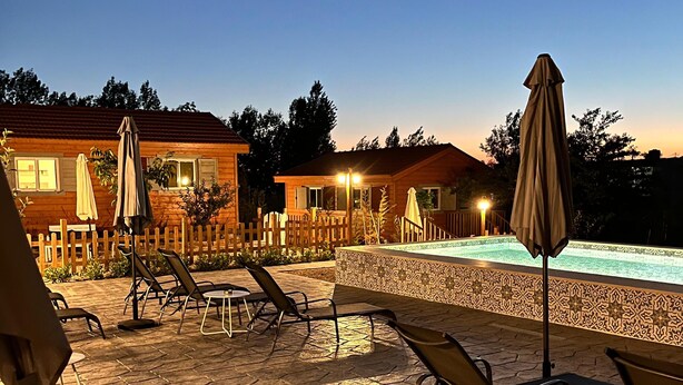 An outdoor patio with lounge chairs and tables, a swimming pool, and wooden cabins in the background. - Beit Mirna Guesthouse & Chalet (Ehden)