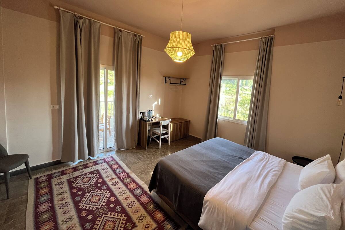 A bedroom with a bed, a desk, and a chair. There are curtains on the windows and a hanging light fixture.