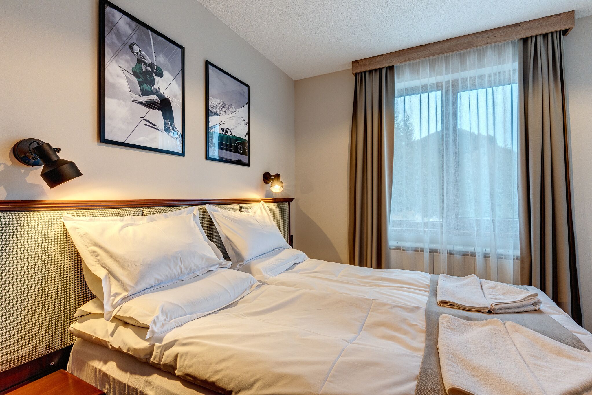 Habitación de hotel con una cama bien hecha, dos cuadros en la pared, una lámpara de noche y un ventanal con cortinas.