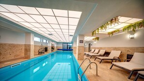Una piscina interior con agua azul cristalina, paredes con azulejos y un techo con diseño de cuadrícula. Hay sillas de descanso y una pequeña zona de estar con una mesa.