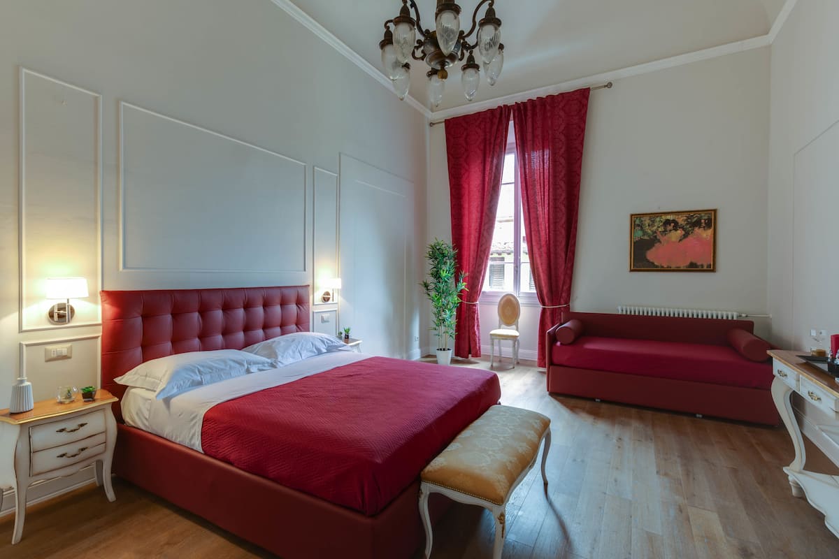 Een slaapkamer met een groot bed, een bankje, een rode bank, een stoel, een kamerplant en een schilderij aan de muur.