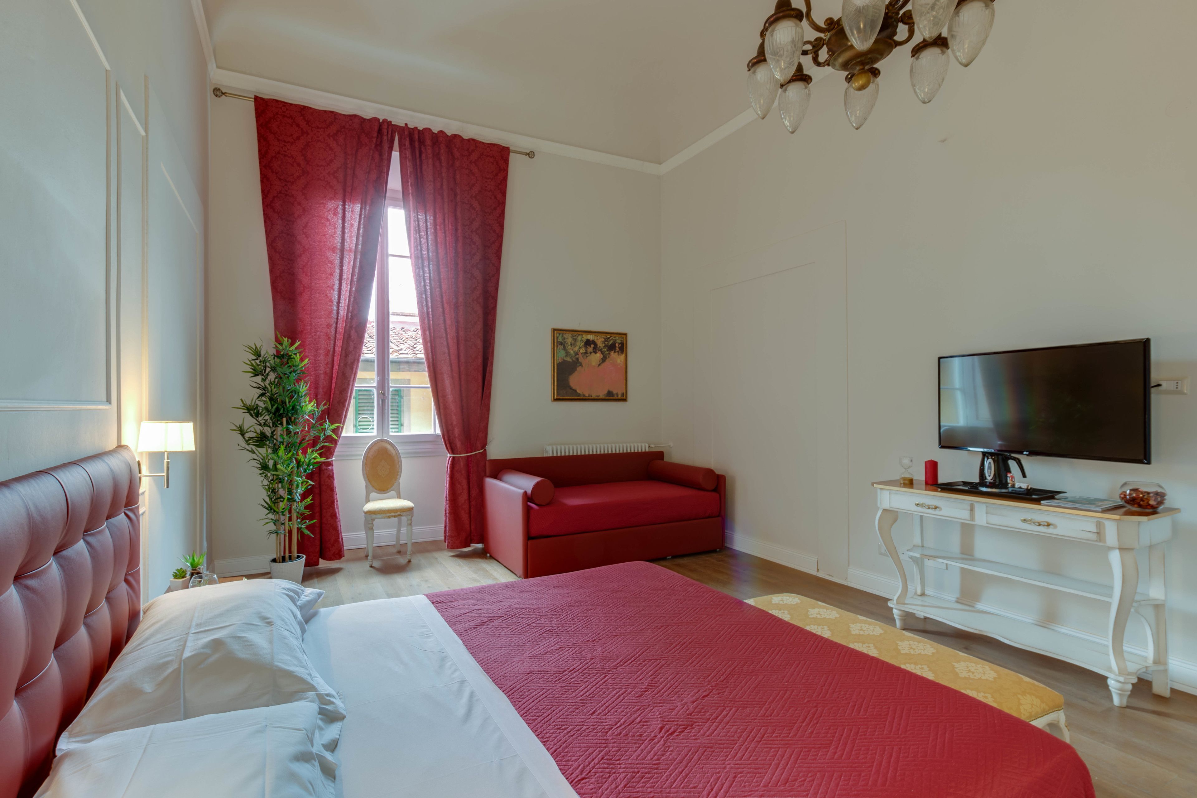 Un dormitorio con una cama grande, un sofá rojo, un televisor sobre un mueble blanco y una ventana con cortinas rojas.
