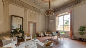 Une pièce spacieuse avec un plafond haut, des décorations élaborées, un lustre et une grande fenêtre avec des rideaux. On y trouve plusieurs fauteuils blancs, une petite table ronde et une plante en pot.