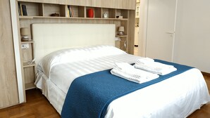 Un lit bien fait, avec une couverture bleue et du linge de lit blanc, une tête de lit en bois et une étagère avec des livres et un vase rouge.